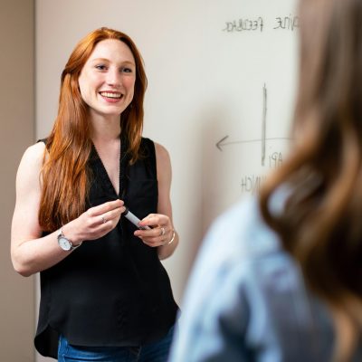 Wir schauen über die Schulter einer Frau und blicken auf eine anderer Frau, die mit einem Marker vor einem Whiteboard steht.