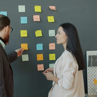 Zwei Personen stehen vor einer Wand mit mehreren Klebezetteln. Der Mann zeigt mit einem Stift auf einen der Zettel.