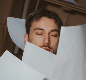 Person mit geschlossenen Augen hinter einem Stapel fallender Papiere
