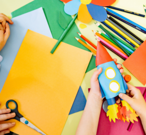Es ist ein Tisch mit Stiften, Tonpapier, Scheren und weiteren Bastelutensilien zu sehen. Zwei Kinderhände halten eine gebastelte Rakete. Weitere Kinderhände sind auf dem Tisch.