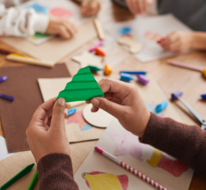Man blickt auf einen bunten Tisch mit Stiften und Papieren und sieht ein paar Kinderhände die daraus etwas basteln.