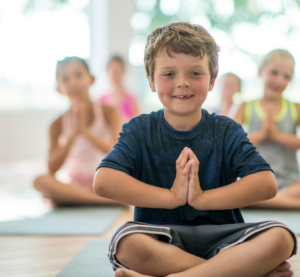 Ein kleiner Junge sitzt im Schneidersitzt. Hinter ihm sind weitere Kinder in der gleichen Yogahaltung zu sehen.