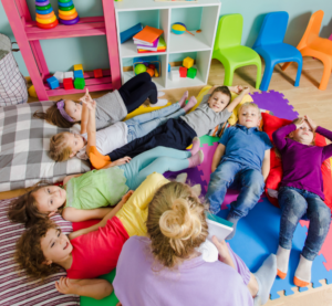 Eine Gruppe von Kindern liegt auf einem Spielteppich, eine Erzieherin sitzt dabei.