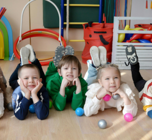 5 Kindergartenkinder liegen auf dem Bauch, haben den Kopf aufgestützt und schauen in die Kamera
