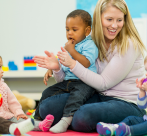Eine Erzieherin sittzt auf dem Boden und klatscht. Sie hat einen kleinen Jungen auf dem Schoß. Neben ihr sitzen noch andere Kinder und alle machen etwas anderes.