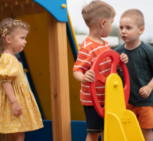 Zwei kleine Jungs streiten um das Steuer eines Spielplatzbootes. Eine kleines Mädchen Beobachtet sie.