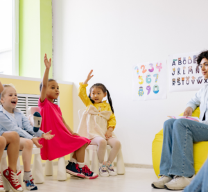 Links sitzen von Kinder im Halbkreis, vier von ihnen heben die Hand. Rechts sitzen zwei Erzieherinnen und halten ein Dokument hoch.