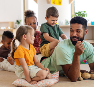 Ein Mann liegt in der Kita auf dem Boden. Drei Kinder sitzen auf seinem Rücken während er mit einem kleinen Mädchen spricht, dass neben ihm sitzt.