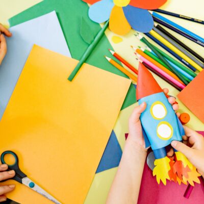 Es ist ein Tisch mit Stiften, Tonpapier, Scheren und weiteren Bastelutensilien zu sehen. Zwei Kinderhände halten eine gebastelte Rakete. Weitere Kinderhände sind auf dem Tisch.