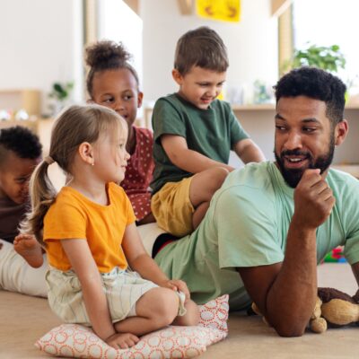 Ein Mann liegt in der Kita auf dem Boden. Drei Kinder sitzen auf seinem Rücken während er mit einem kleinen Mädchen spricht, dass neben ihm sitzt.