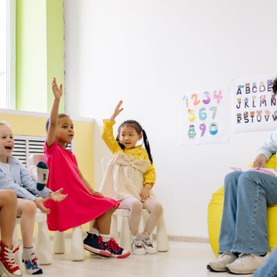 Links sitzen von Kinder im Halbkreis, vier von ihnen heben die Hand. Rechts sitzen zwei Erzieherinnen und halten ein Dokument hoch.