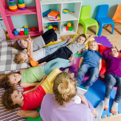 Eine Gruppe von Kindern liegt auf einem Spielteppich, eine Erzieherin sitzt dabei.