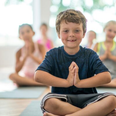Ein kleiner Junge sitzt im Schneidersitzt. Hinter ihm sind weitere Kinder in der gleichen Yogahaltung zu sehen.