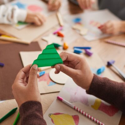 Man blickt auf einen bunten Tisch mit Stiften und Papieren und sieht ein paar Kinderhände die daraus etwas basteln.