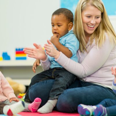 Eine Erzieherin sittzt auf dem Boden und klatscht. Sie hat einen kleinen Jungen auf dem Schoß. Neben ihr sitzen noch andere Kinder und alle machen etwas anderes.