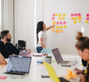Eine Frau steht an einer Wand und zeigt auf die Klebezettel, die dort hängen. Davor steht ein Tisch, an dem zwei Frauen und zwei Männer mit Laptop sitzen und sich Notizen machen.