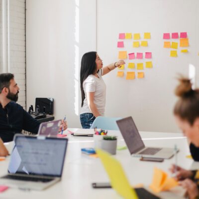 Eine Frau steht an einer Wand und zeigt auf die Klebezettel, die dort hängen. Davor steht ein Tisch, an dem zwei Frauen und zwei Männer mit Laptop sitzen und sich Notizen machen.
