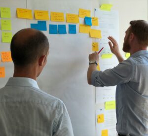 Zwei Männer stehen vor eine Whiteboard mit Klebezetteln. Sie diskutieren und der Eine Mann versetzt Zettel.