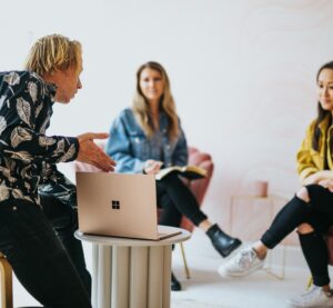 Drei Jugendliche sitzen zusammen. Ein junger Mann hat vor sich einen kleinen Tisch mit Laptop und zeigt den beiden jungen Frauen darauf etwas.