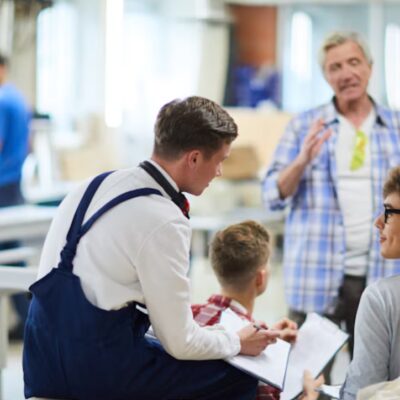 Drei männliche Jugendliche in blauen Latzhosen sitzen in einer Werkstatt zusammen. Vor Ihm steht ein älterer Arbeiter steht vor ihnen und erklärt etwas.