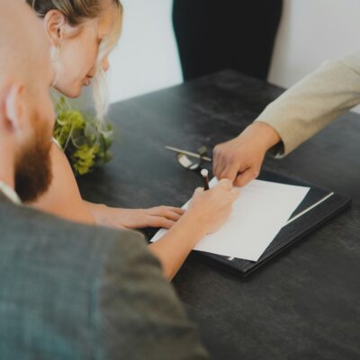 Eine Frau sitzt an einem Konferenztisch und schreibt etwas auf. Jemand sitzt ihr gegenüber und zeigt etwas auf dem Blatt Papier. Neben der Frau sitzt noch ein Mann, dem wir über die Schulter schauen.