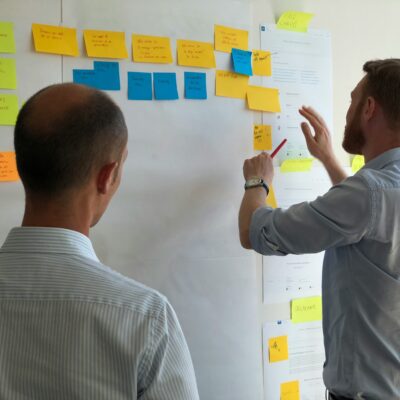 Zwei Männer stehen vor eine Whiteboard mit Klebezetteln. Sie diskutieren und der Eine Mann versetzt Zettel.