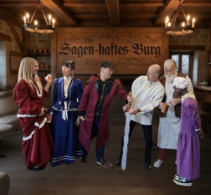 Verschiedene Personen in mittelalterlichen Kostümen stehen in einem hölzernen Raum. Hinter ihnen prangt an der Wand "Sagen-haftes Burg".